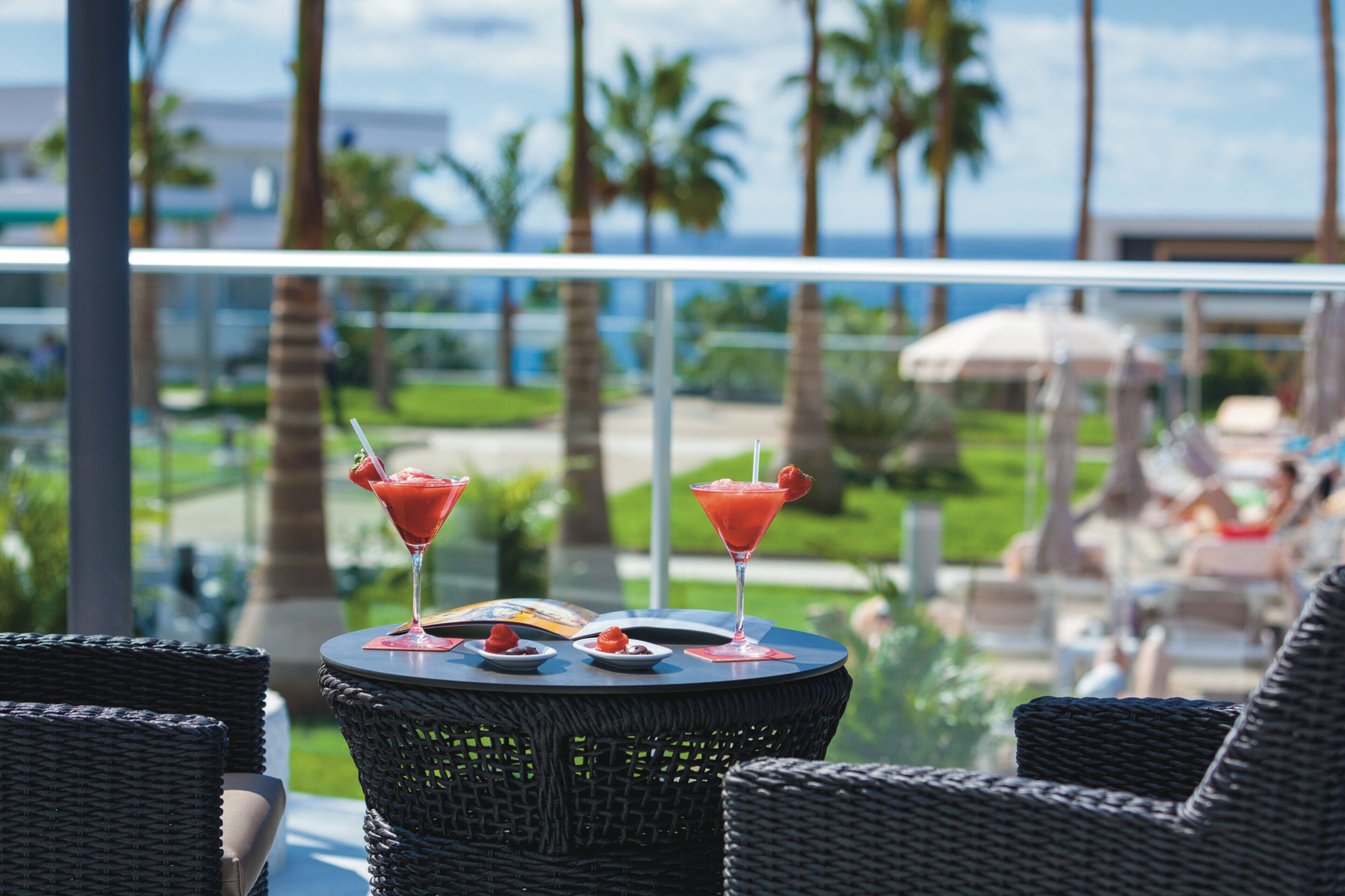 A balcony with wicker chairs, a table with two red cocktails and desserts, and a view of palm trees and a pool.