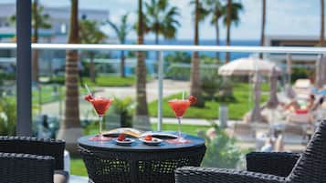 A balcony with wicker chairs, a table with two red cocktails and desserts, and a view of palm trees and a pool.
