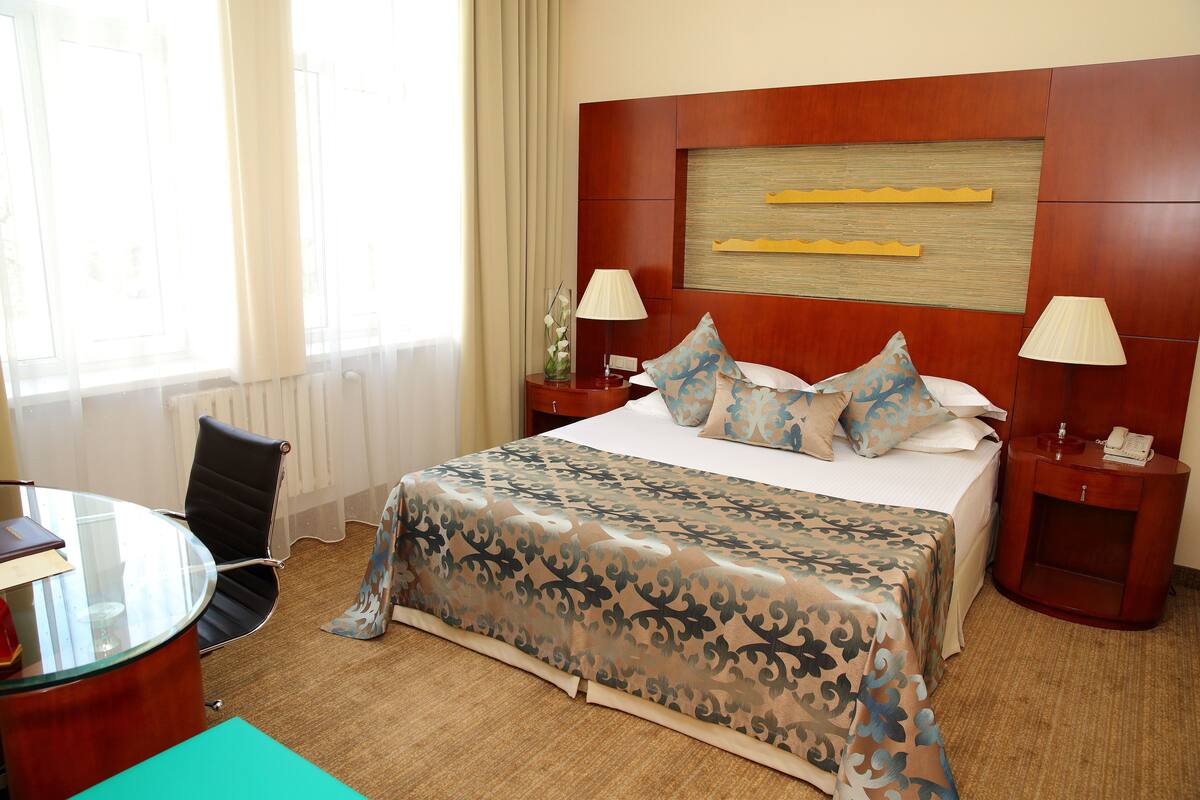 Habitación de hotel con una cama grande, dos mesitas de noche con lámparas, un cabecero de madera, un escritorio de cristal con una silla y vistas al exterior.