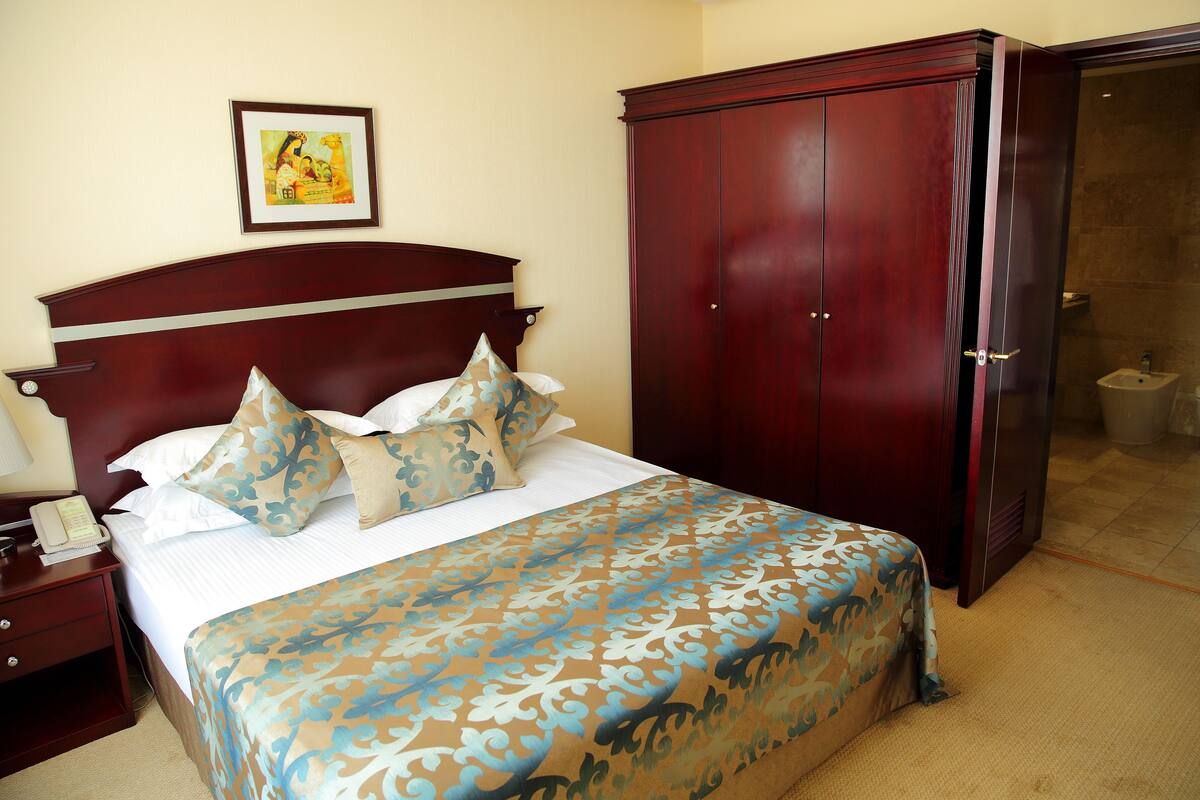 Una cama bien tendida con una colcha estampada, una mesita de noche de madera con un teléfono, un cuadro en la pared y un armario de madera oscura.