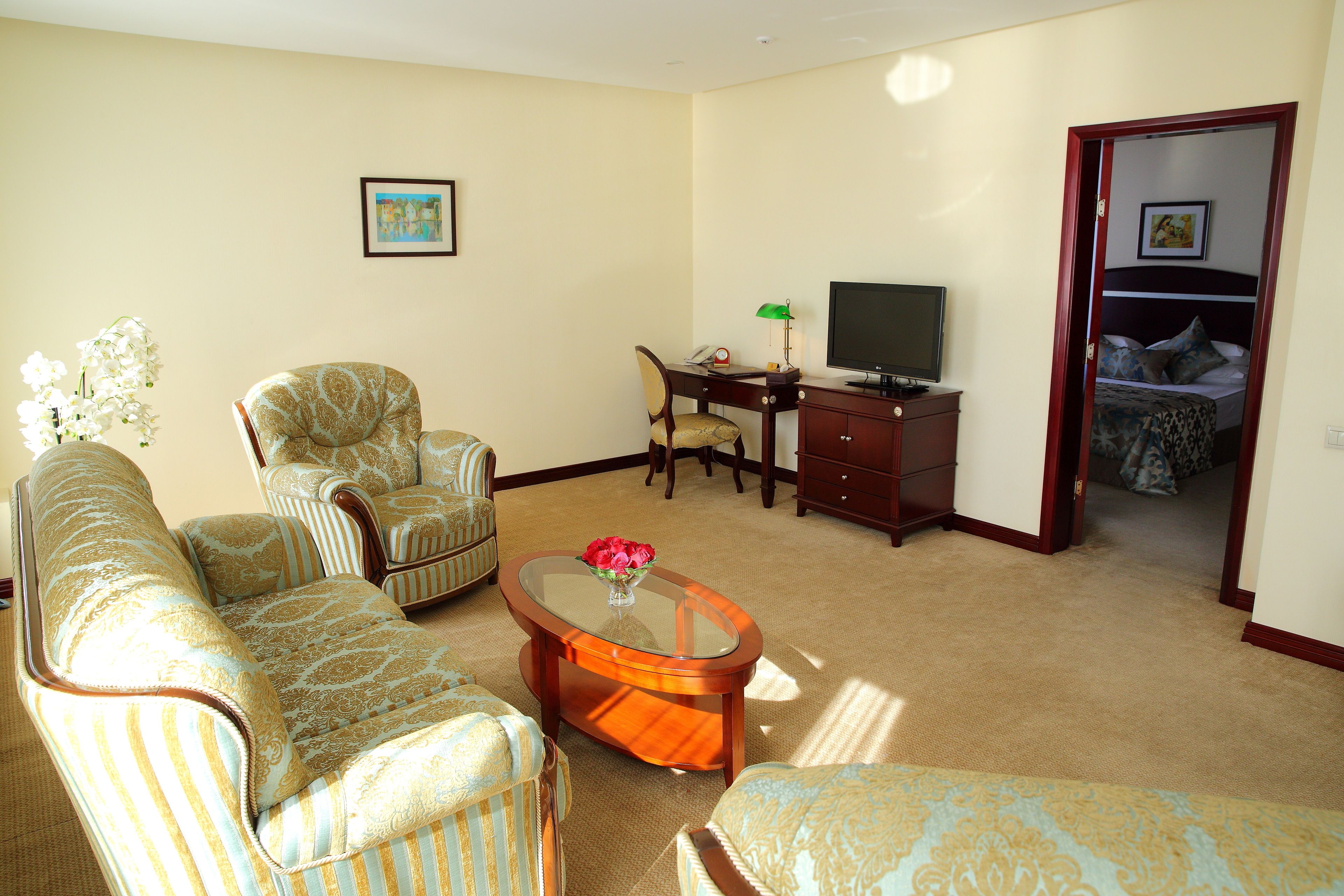 Una sala de estar con sofá, sillón y una mesa de centro de vidrio. Hay un televisor sobre un mueble de madera y una cama al fondo.