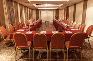 Uma sala de conferência com uma mesa comprida, toalha vermelha, cadeiras douradas e piso coberto de carpete.