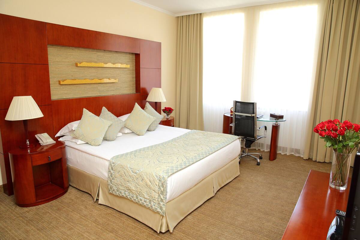 Habitación de hotel con una cama grande, dos mesitas de noche con lámparas, un escritorio con silla, un televisor y un jarrón de flores.