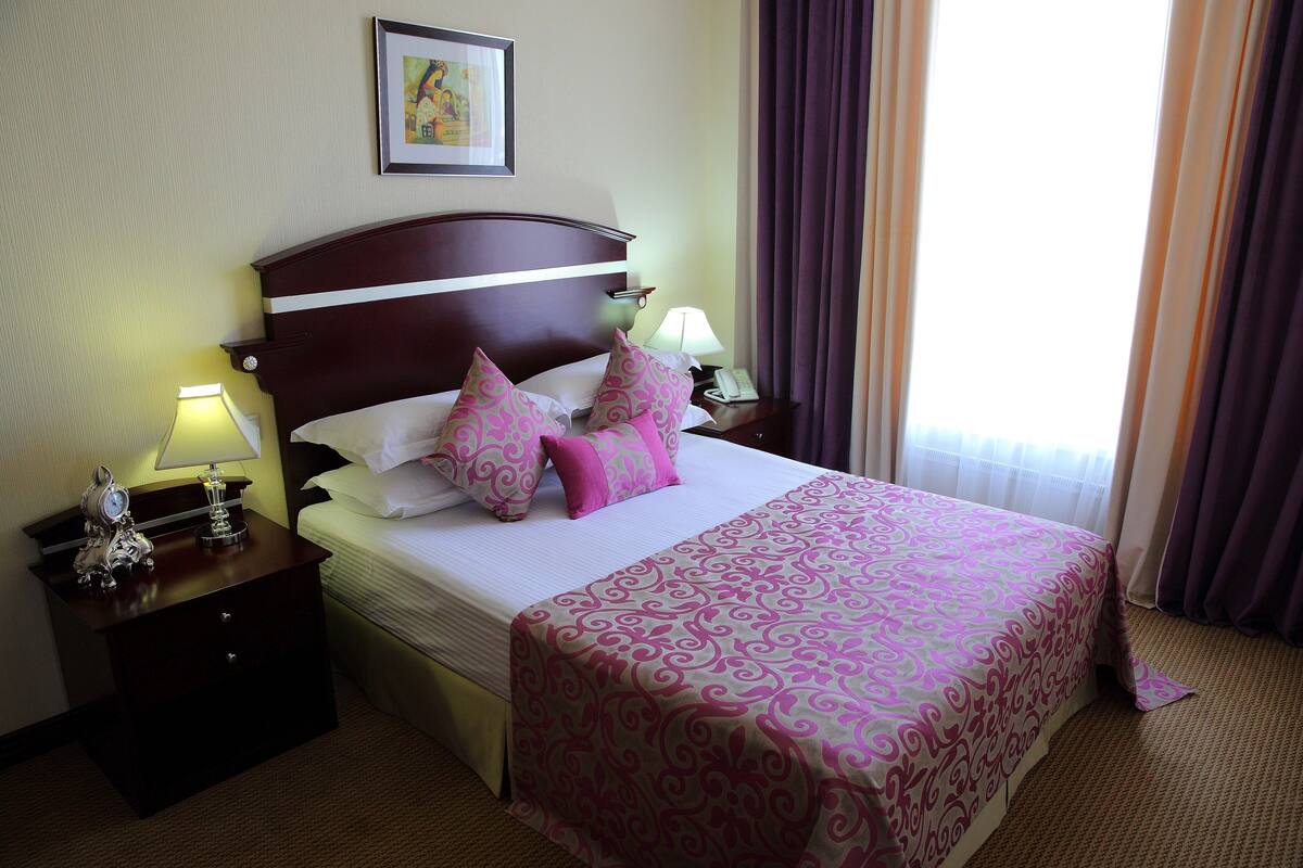 Una habitación de hotel con una cama grande, una mesita de noche con lámpara, un cuadro enmarcado en la pared y una ventana con cortinas.