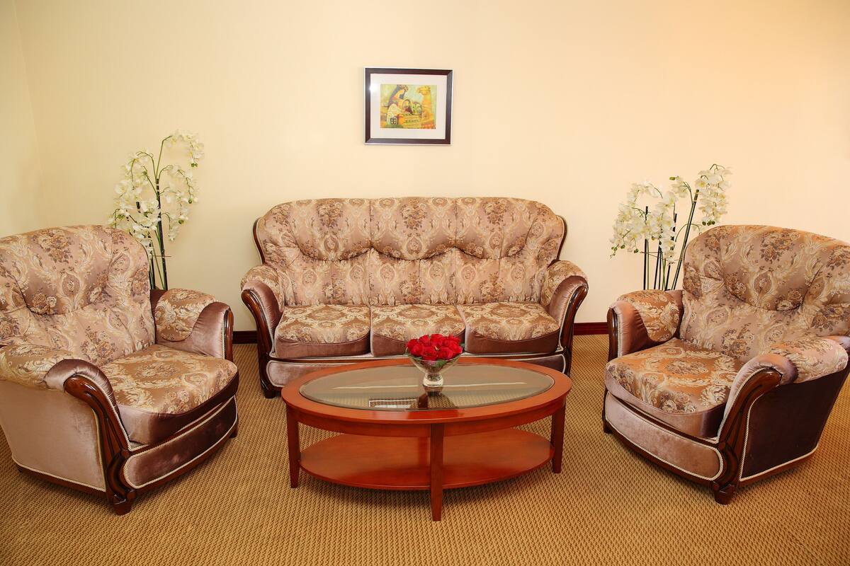 Una sala de estar con un sofá estampado, dos sillones, una mesa de centro de vidrio con un jarrón de flores y un cuadro en la pared.