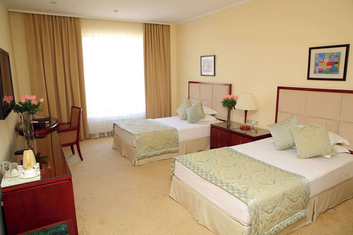 Una habitación de hotel con dos camas, un escritorio, una silla, una mesita de noche y una ventana con cortinas.