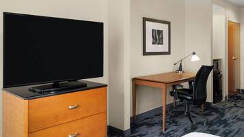 Et hotelværelse med et fladskærms-tv på en trækommode, et skrivebord med stol, et billede i ramme på væggen, og en dør der fører til et andet rum.