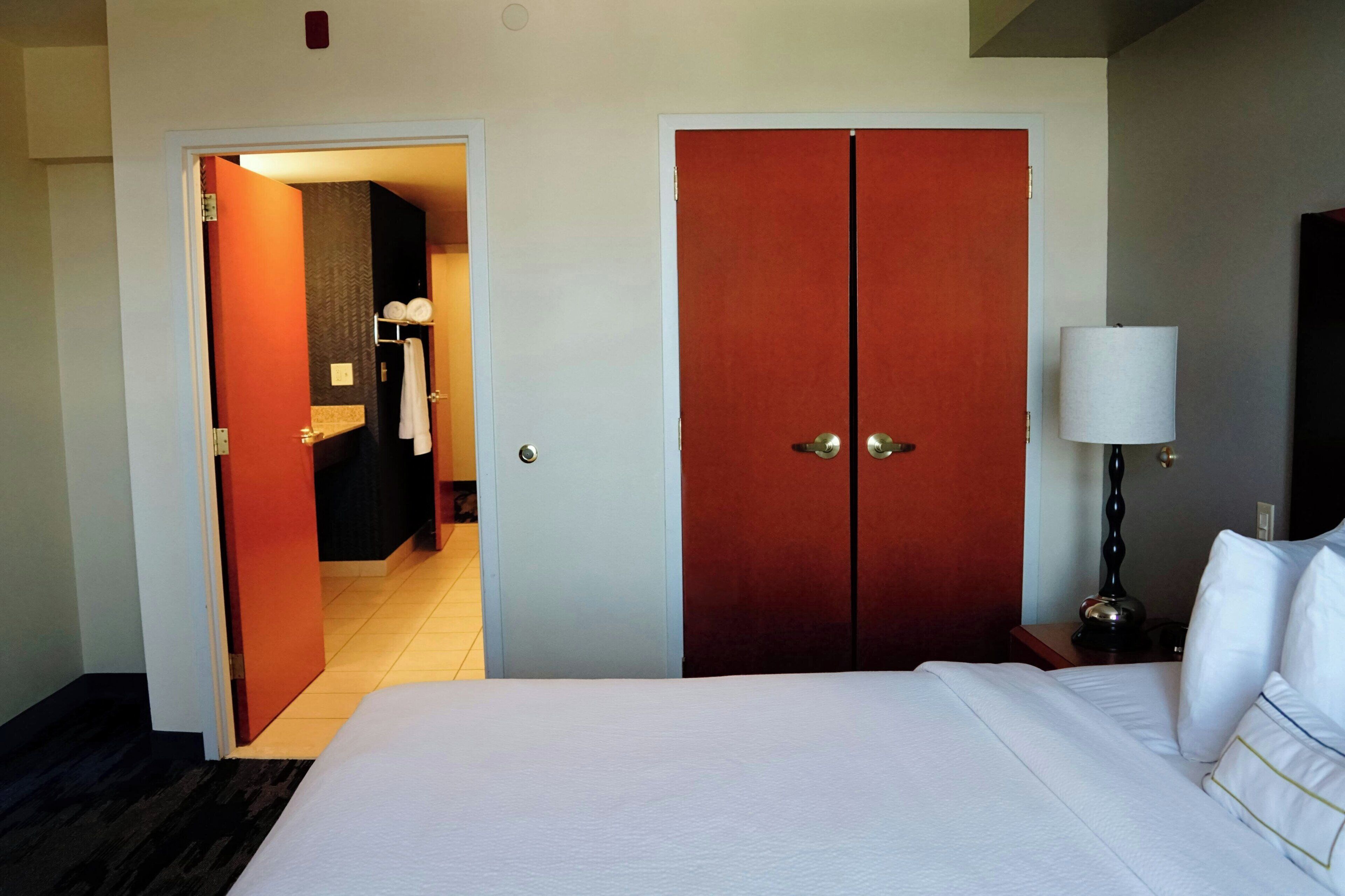 Una habitación de hotel con una cama grande, un armario, una mesita de noche con lámpara y un baño visible a través de una puerta abierta.