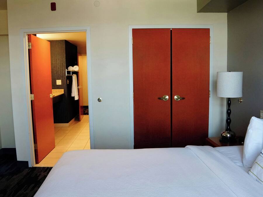 Une chambre d’hôtel avec un grand lit, une armoire, une table de chevet avec une lampe et une salle de bain visible à travers une porte ouverte.