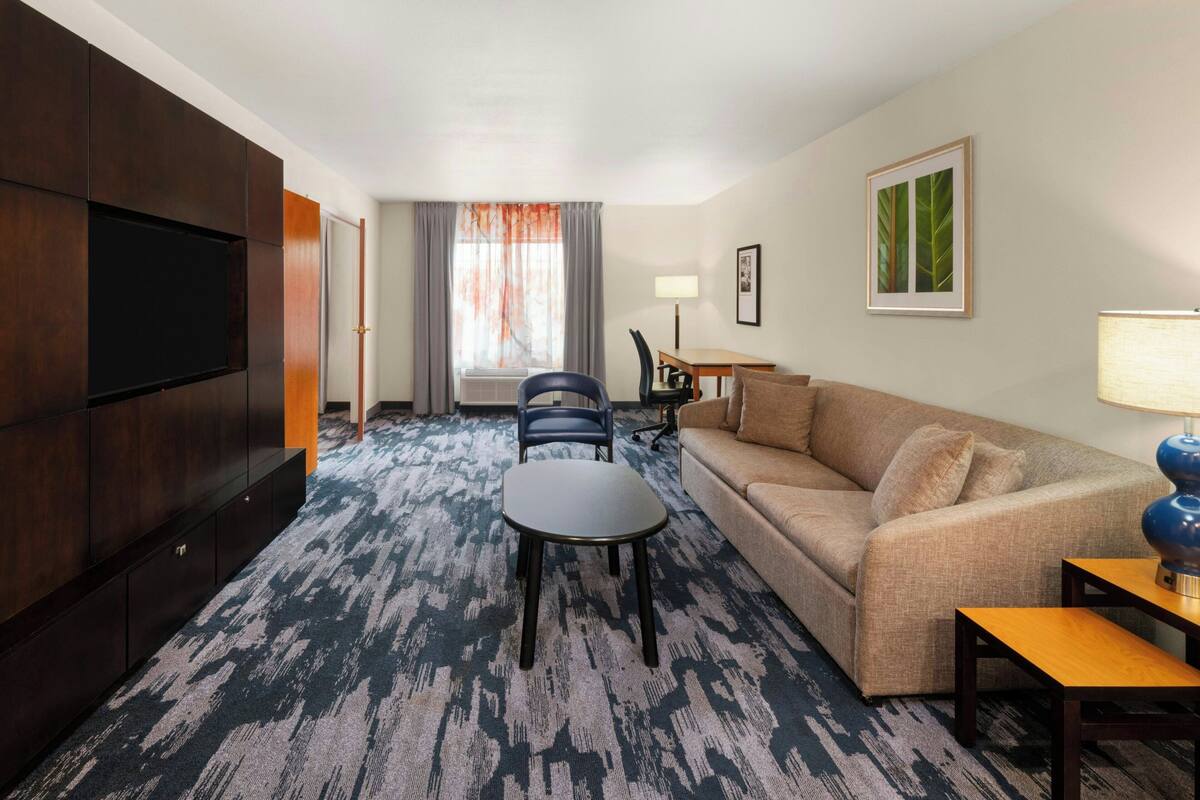 Habitació d'hotel amb un sofà, una taula petita, un televisor, una làmpada i una finestra amb cortines.