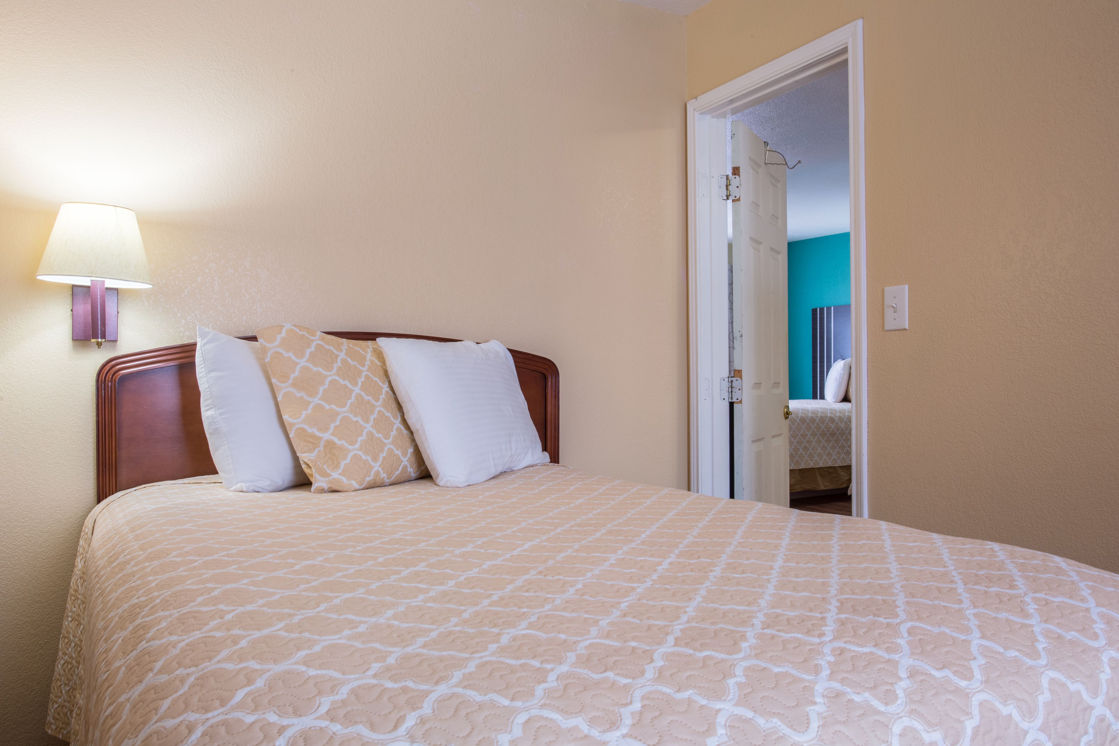 Quarto com cama, mesa de cabeceira, luminária e porta que dá para outro espaço.