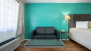A hotel room with a teal wall, a black sofa, a small table, a bed with a patterned bedspread, a bedside lamp, and a window with a curtain. - Pinn Road Inn and Suites (San Antonio)