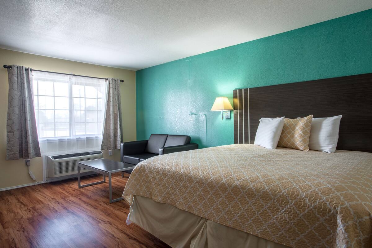 A hotel room with a large bed, a desk, a chair, a window with curtains, and a wall-mounted lamp.