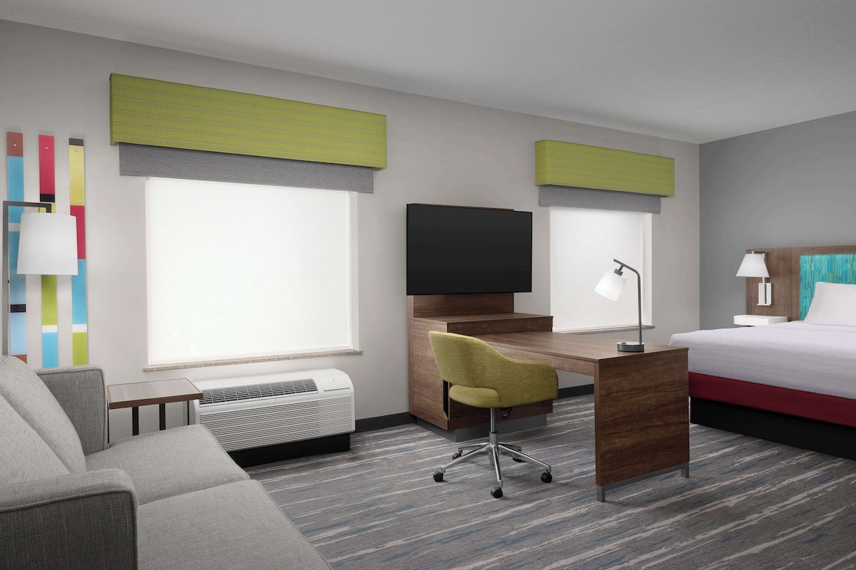 Ein Hotelzimmer mit Bett, Schreibtisch mit Stuhl, Fernseher und einem Fenster mit Jalousien.