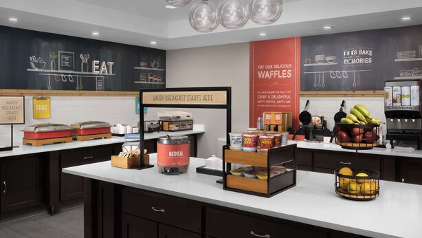 A breakfast area with a chalkboard menu, a sign indicating breakfast starts here, and a variety of food items like waffles, oatmeal, and fruits.