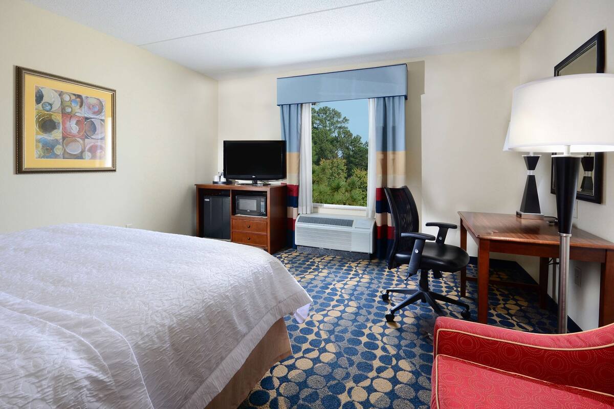 Ein Hotelzimmer mit Bett, Schreibtisch, Stuhl, Fernseher und einem Fenster mit Vorhängen.