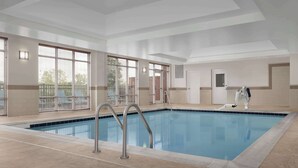A large indoor swimming pool with clear blue water, surrounded by beige tiled flooring and walls, with large windows allowing natural light.