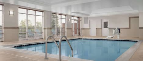 A large indoor swimming pool with clear blue water, surrounded by beige tiled flooring and walls, with large windows allowing natural light.