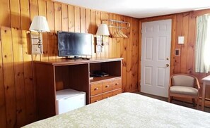 A room with wooden walls, a bed, a television on a wooden stand, a white door, and a chair.