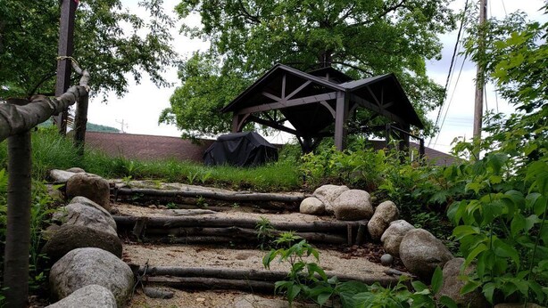 Une structure rustique en bois, dotée d’un toit et entourée de végétation naturelle et de rochers.