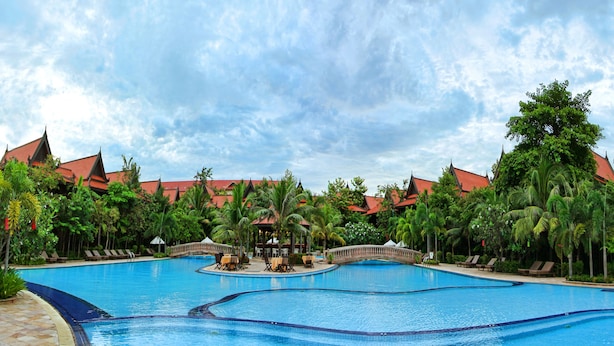 A large outdoor swimming pool with multiple sections, surrounded by palm trees and a building with red roofs. - Sokhalay Angkor Villa Resort (Siem Reap)