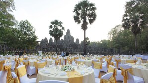 An outdoor event space with round tables set for dining, surrounded by white chairs with gold sashes, and palm trees in the background. - Sokhalay Angkor Villa Resort (Siem Reap)