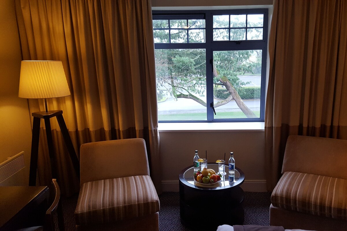 A hotel room with two upholstered chairs, a round table with a fruit bowl and bottles, a lamp, and a window with curtains.