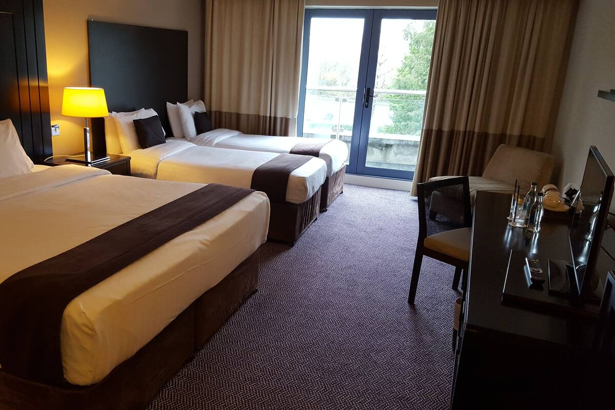 A hotel room with two large beds, a desk with a chair, a lamp, and a view of the outdoors through a large window.