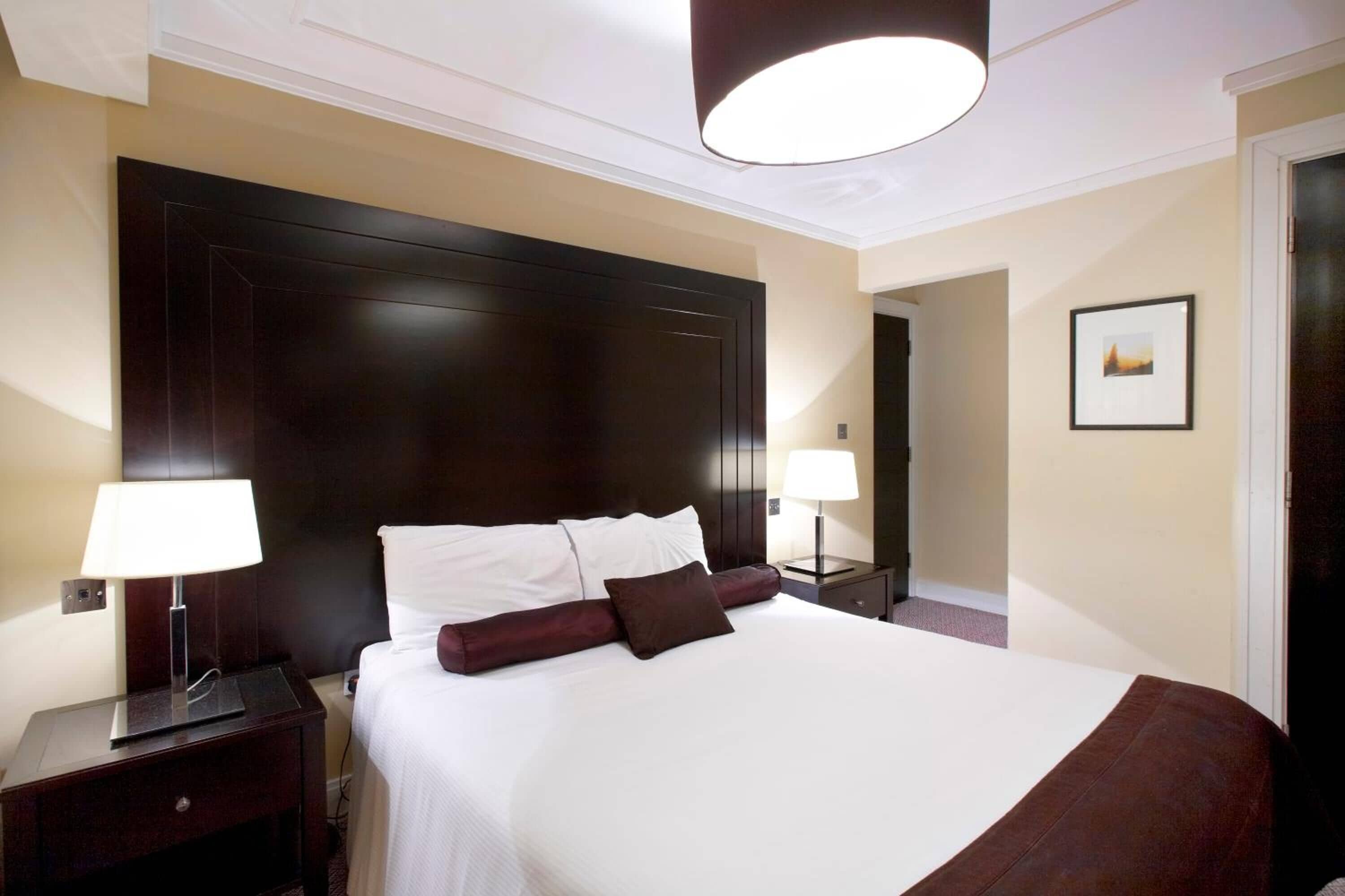 Habitación de hotel con una cama grande, dos mesitas de noche con lámparas, un cabecero de madera oscura y un cuadro en la pared.