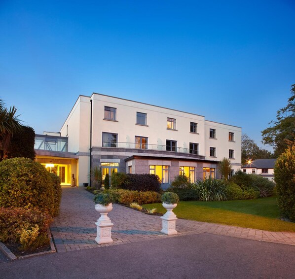 Un hotel moderno con fachada simétrica, numerosas ventanas y un jardín bien cuidado con senderos y urnas decorativas.