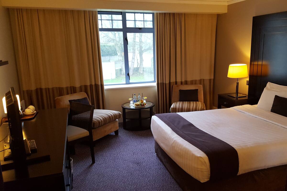 A hotel room with a large bed, a desk, a chair, a small table with a bottle and a glass, and a window with curtains.