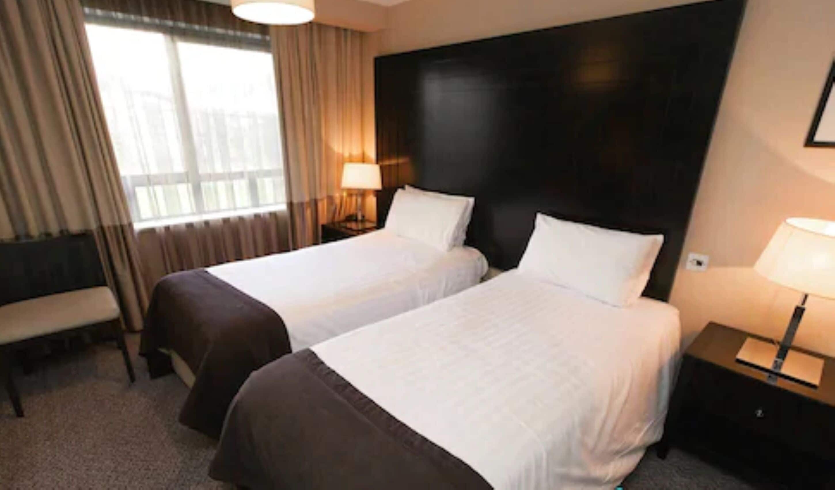 Habitación de hotel con dos camas, un banco, una mesita de noche con lámpara y un ventanal con cortinas.