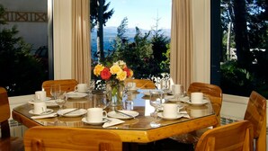 Una mesa de comedor dispuesta para seis personas, con platos, tazas y cubiertos blancos. Alrededor, sillas de madera. Un jarrón con flores amarillas y rojas está sobre la mesa.