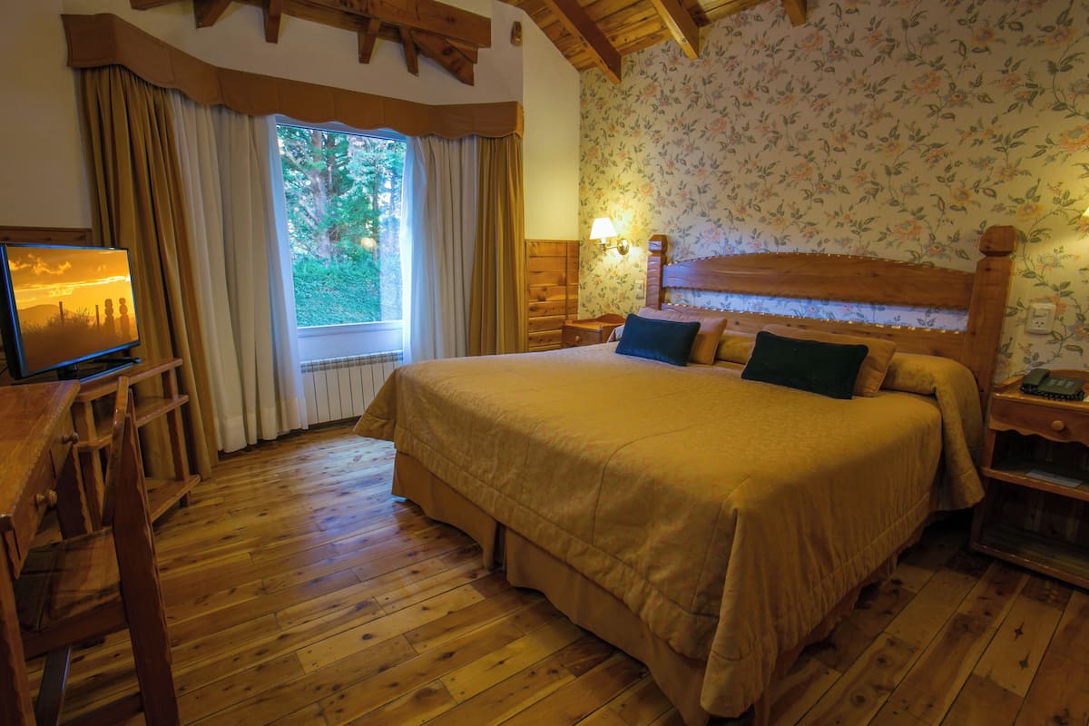 A wooden-framed bedroom with a large bed, a television on a wooden table, and a window with curtains.