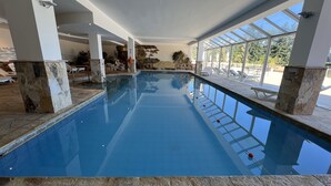 Una piscina interior con paredes de vidrio transparente, pilares de piedra y sillones de descanso afuera.