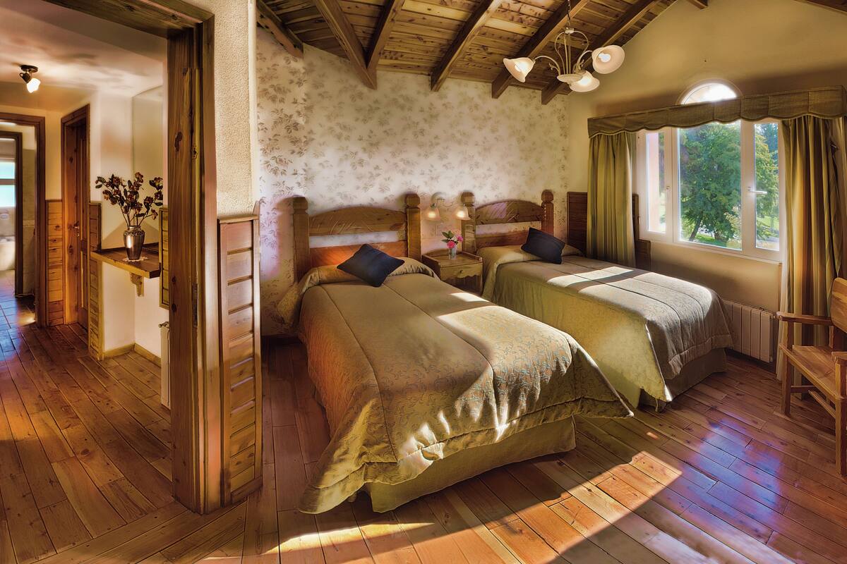 A room with two wooden beds, a wooden bench, a window with curtains, and a wooden ceiling.