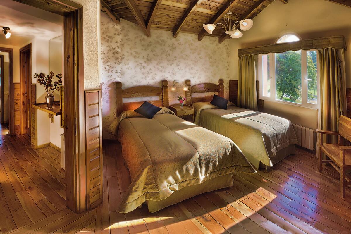 A room with two wooden beds, a wooden bench, a window with curtains, and a wooden ceiling.