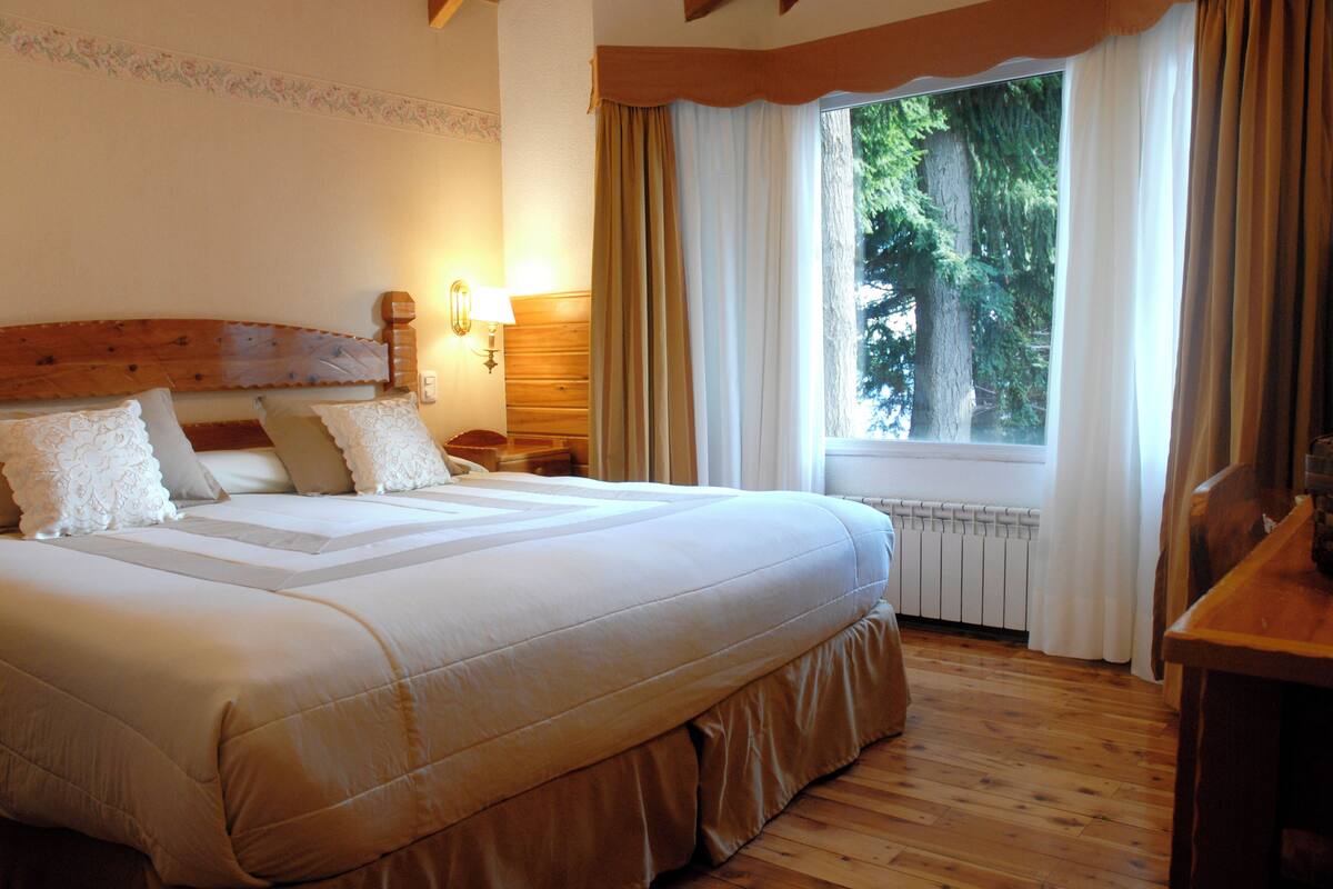 A large bed with a wooden headboard, a nightstand, a lamp, and a window with curtains.