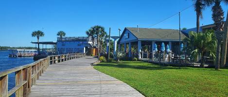 A wooden pier leading to a building with outdoor seating and palm trees.