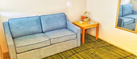 A room with a blue sofa, a small wooden table with a basket of flowers, a framed picture on the wall, and a large mirror.