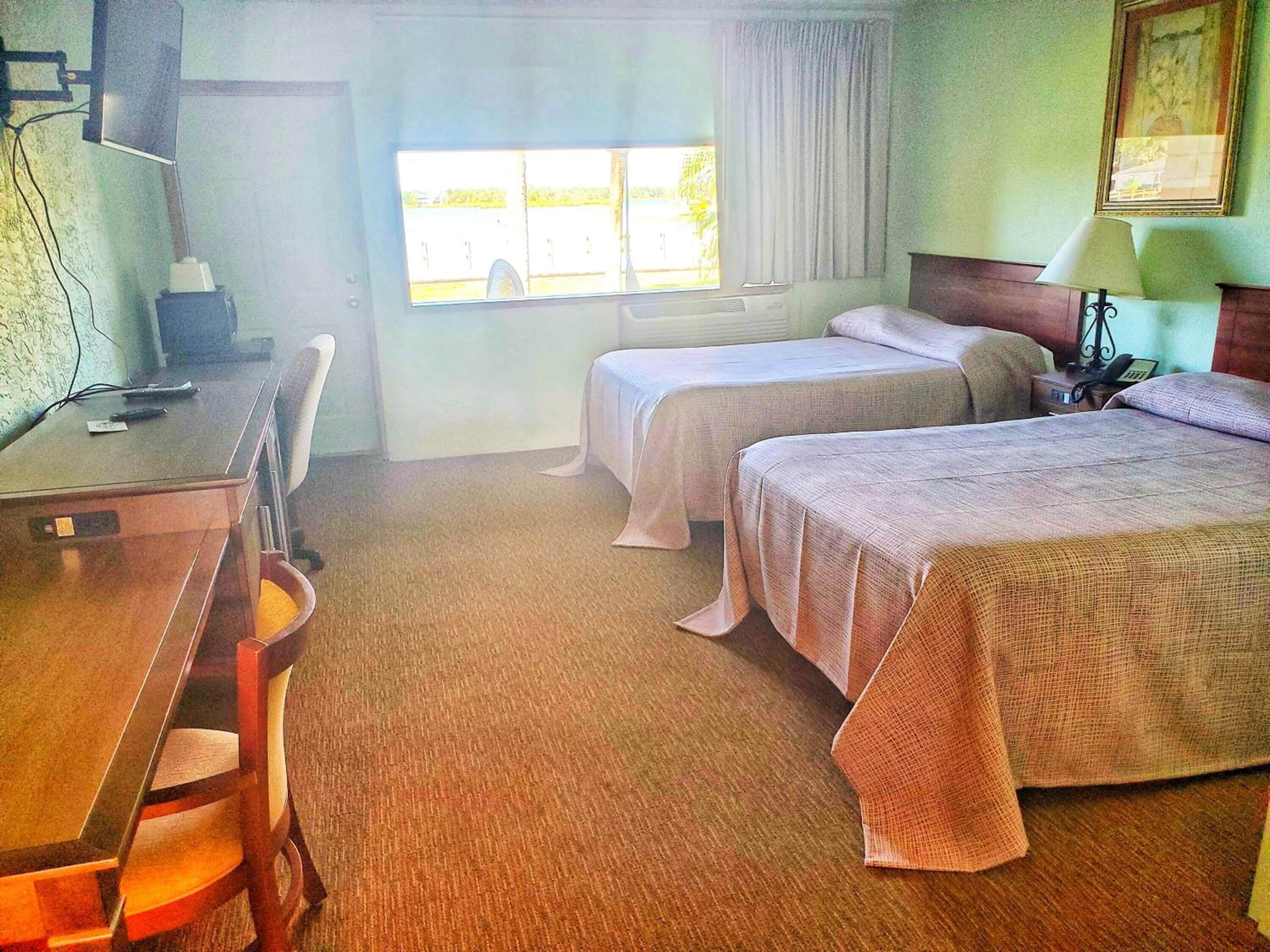 Une chambre d’hôtel avec un bureau, deux lits, une chaise, une fenêtre et une lampe.