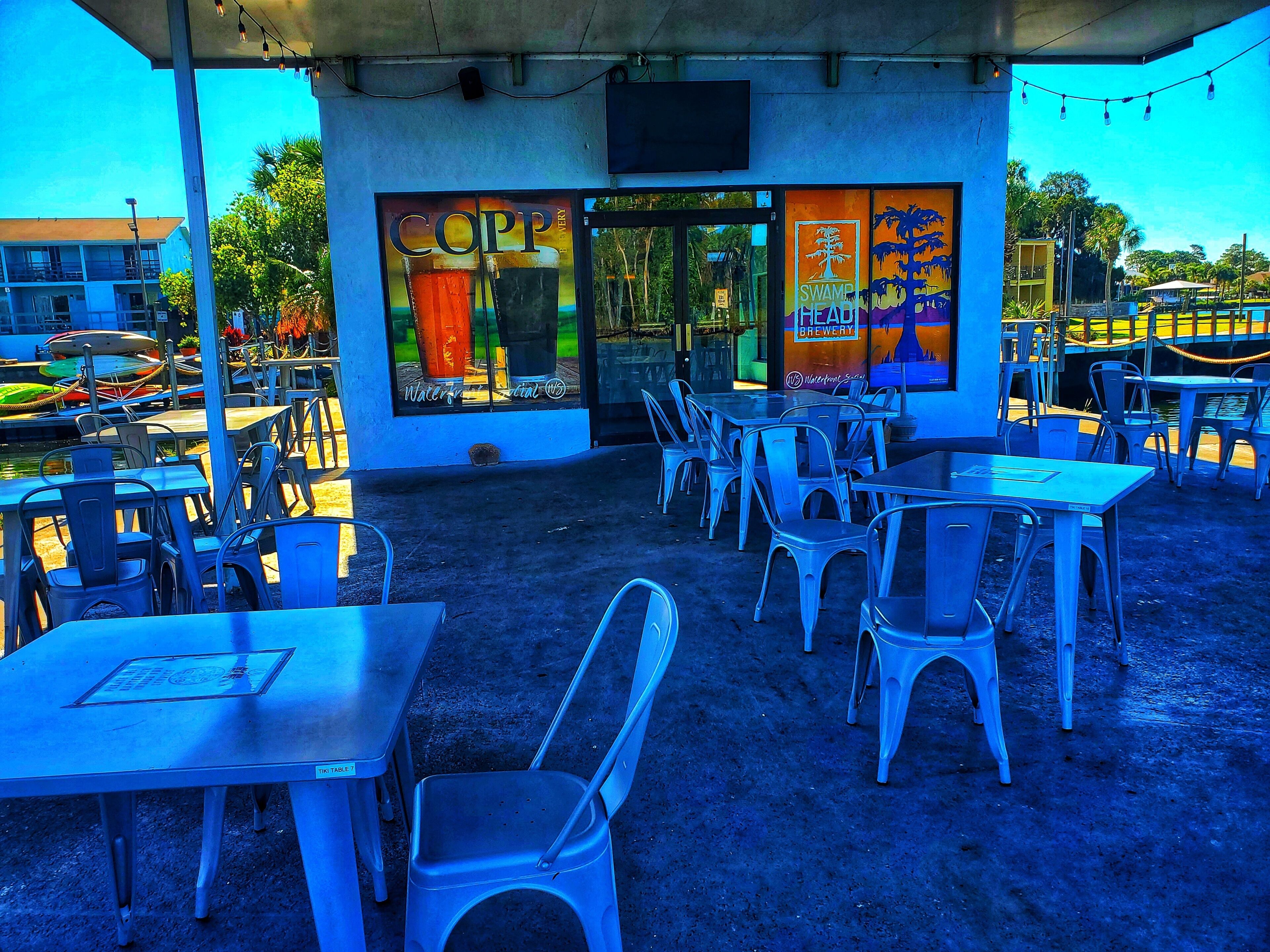 An outdoor seating area with tables and chairs, a covered section with a sign for 'COPP SWAMP BREWERY', and a view of the surrounding area including parked boats and buildings.