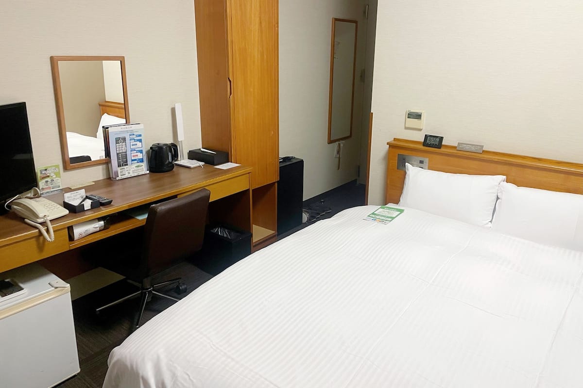 A hotel room with a bed, a desk with a telephone, a mirror, a wardrobe, and a nightstand with a clock.