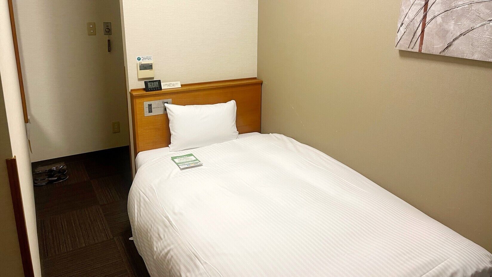 A hotel room with a single bed, a wooden headboard, a white pillow, a bedside table with a clock, and a small sign on the wall.