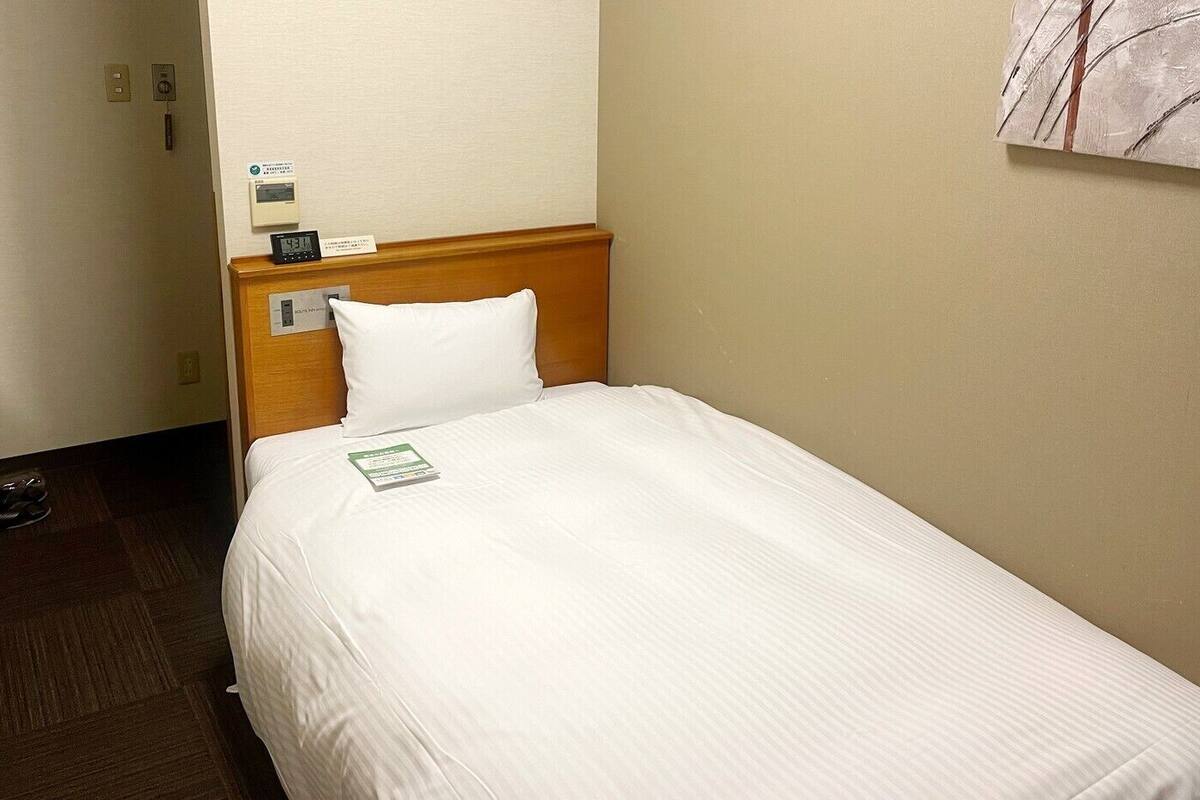 A hotel room with a single bed, a wooden headboard, a white pillow, a bedside table with a clock, and a small sign on the wall.