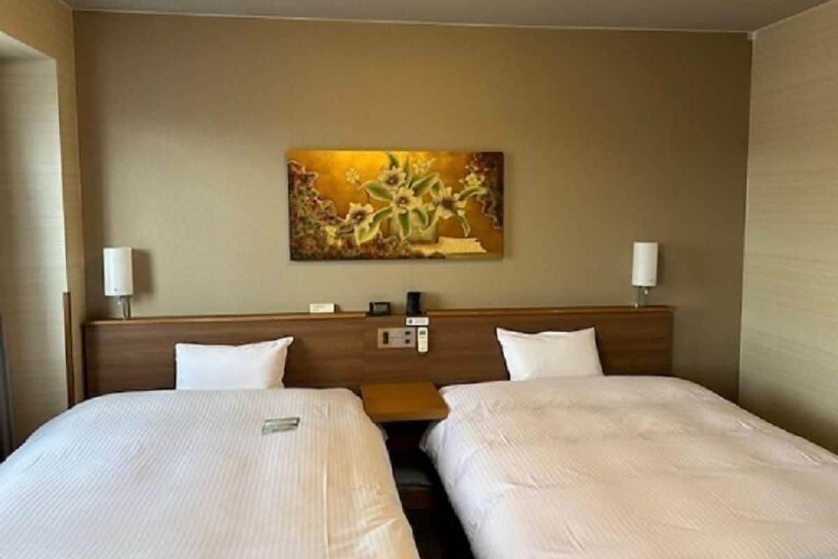 A hotel room with two beds, a painting on the wall, and a nightstand with a lamp.