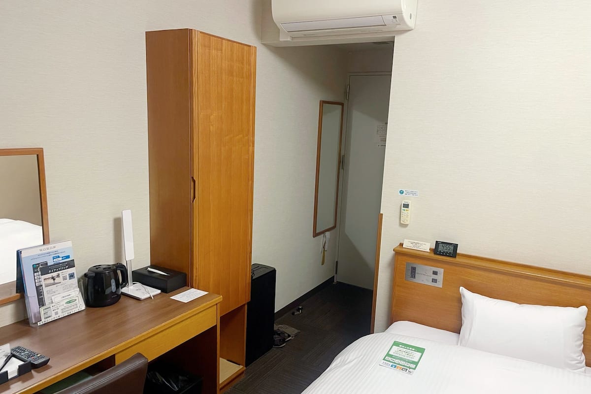 A hotel room with a wooden wardrobe, a desk with a coffee maker, a mirror, a bed with white bedding, and a bedside table.