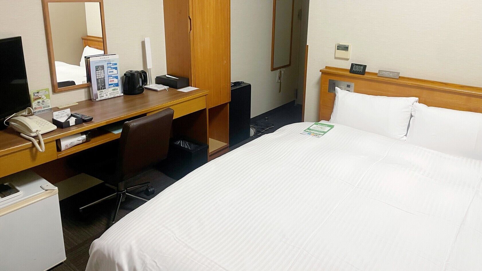 A hotel room with a bed, a desk with a telephone, a chair, a mirror, and a clock.