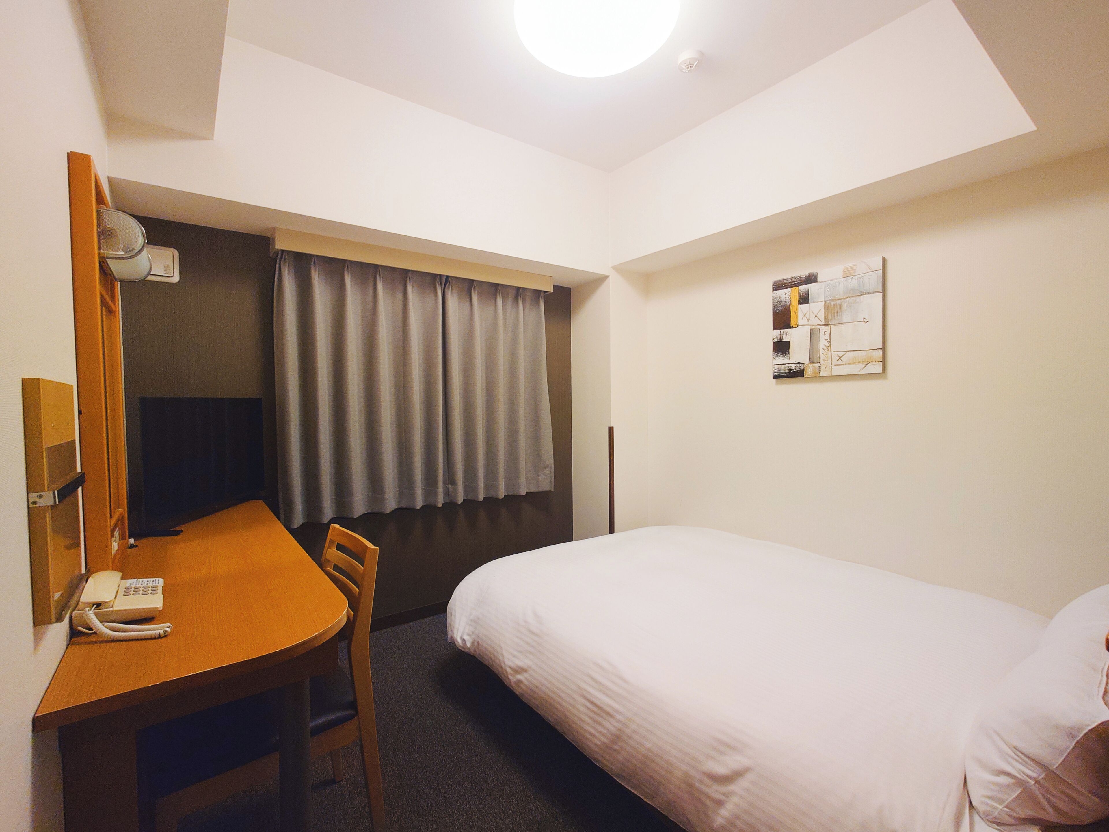 Habitació d'hotel amb un llit, una taula de nit amb telèfon, una cadira, una finestra amb cortines i una obra d'art penjada a la paret.