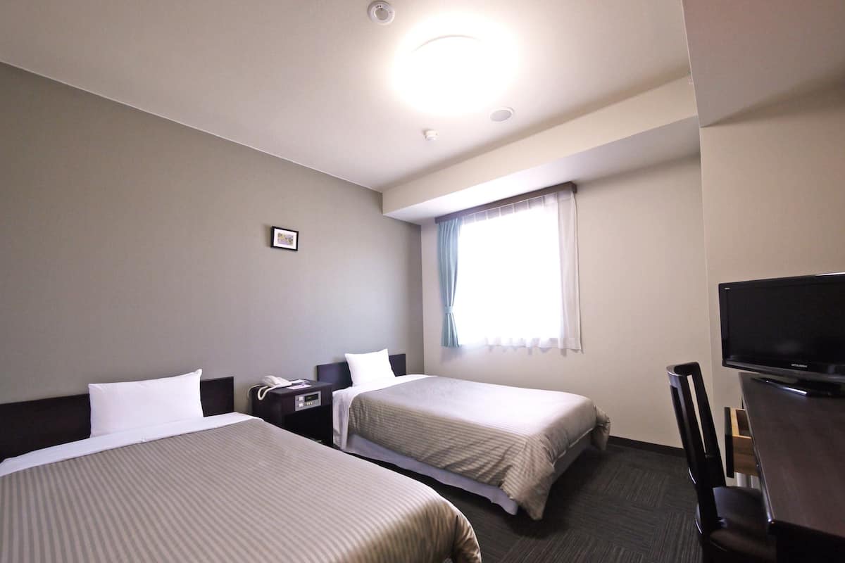 A hotel room with two beds, a desk with a chair, a television, and a window with curtains.